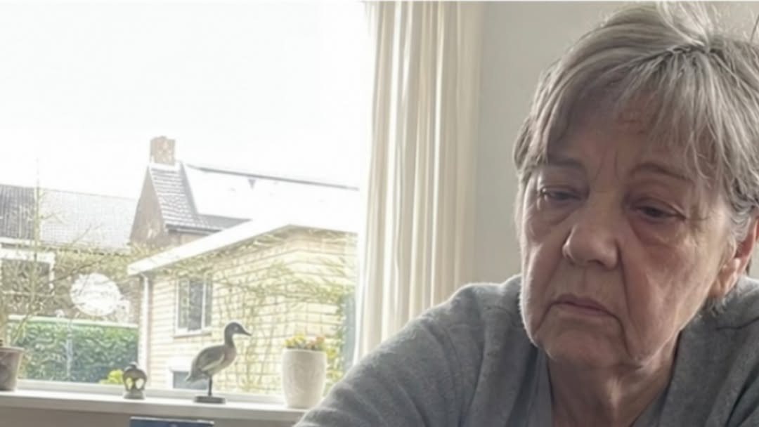 Een vrouw met grijs krullend haar in een beige vest zit thuis in de woonkamer
